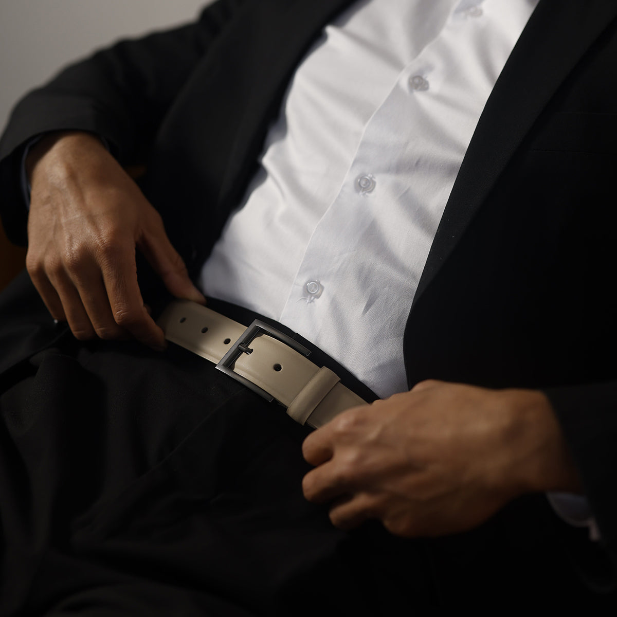 A man dressed in a black suit and white shirt is adjusting a stylish beige Silvano leather belt.