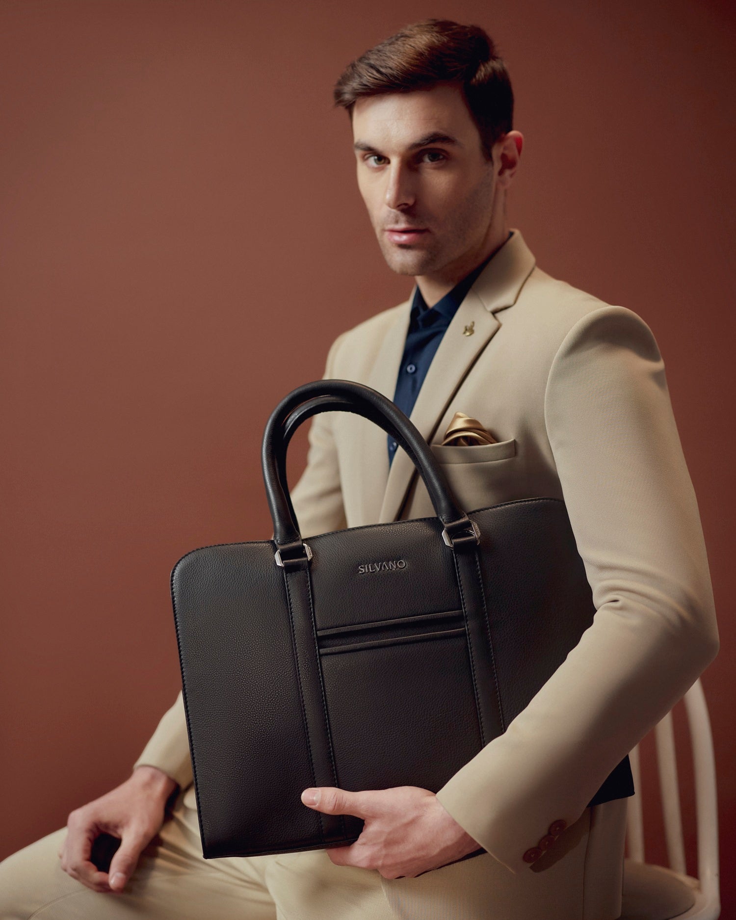 silvano mayfair laptop bag - black leather office bag held by man in beige suit against brown background