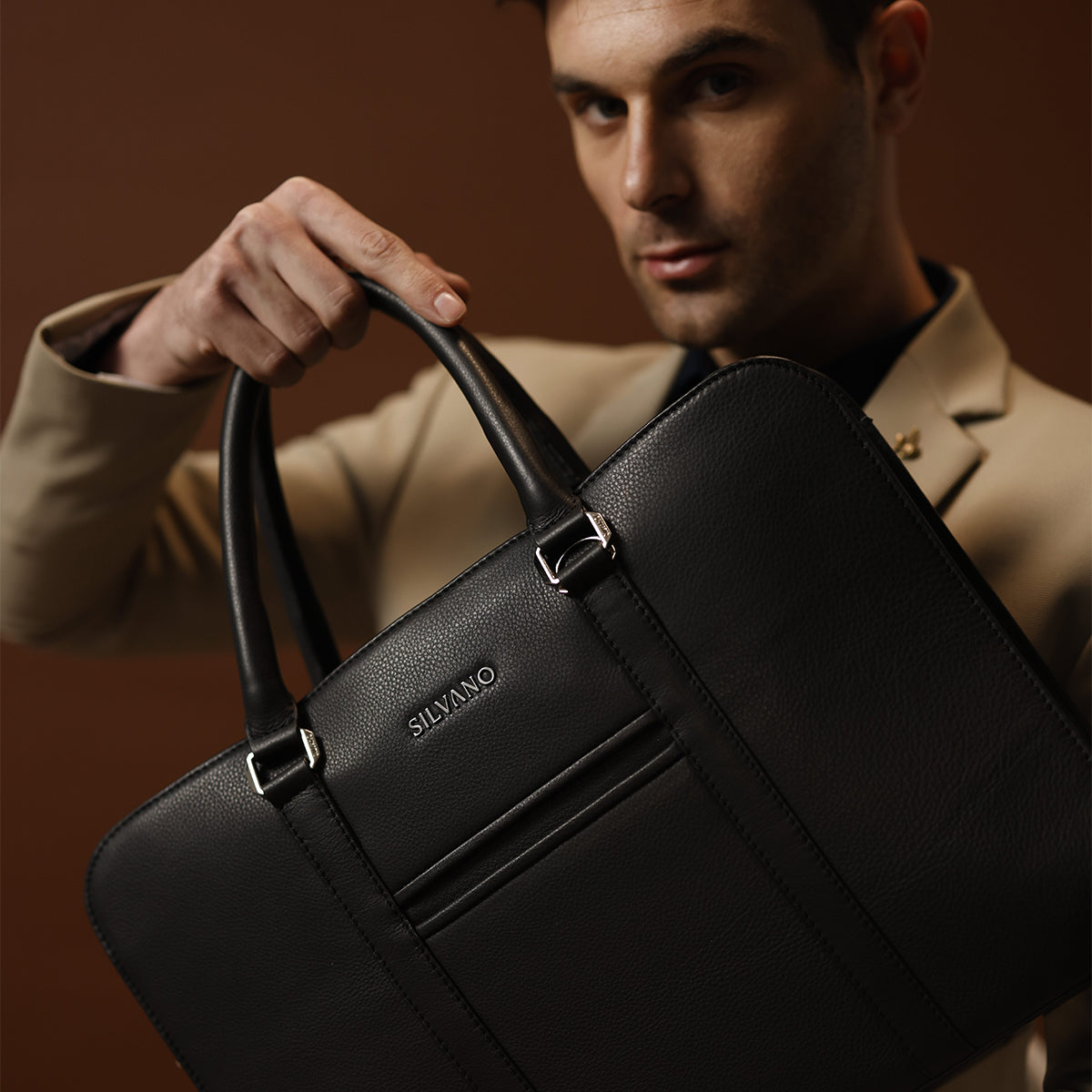 A stylish man in a beige blazer confidently holds up a sleek black Silvano leather laptop bag, showcasing its premium design and dual-handle structure.
