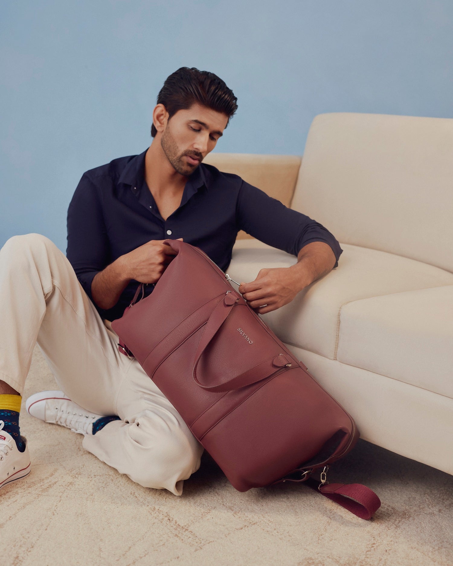 silvano gear grip weekender duffle bag - maroon leather duffle bag held by man sitting beside a beige sofa